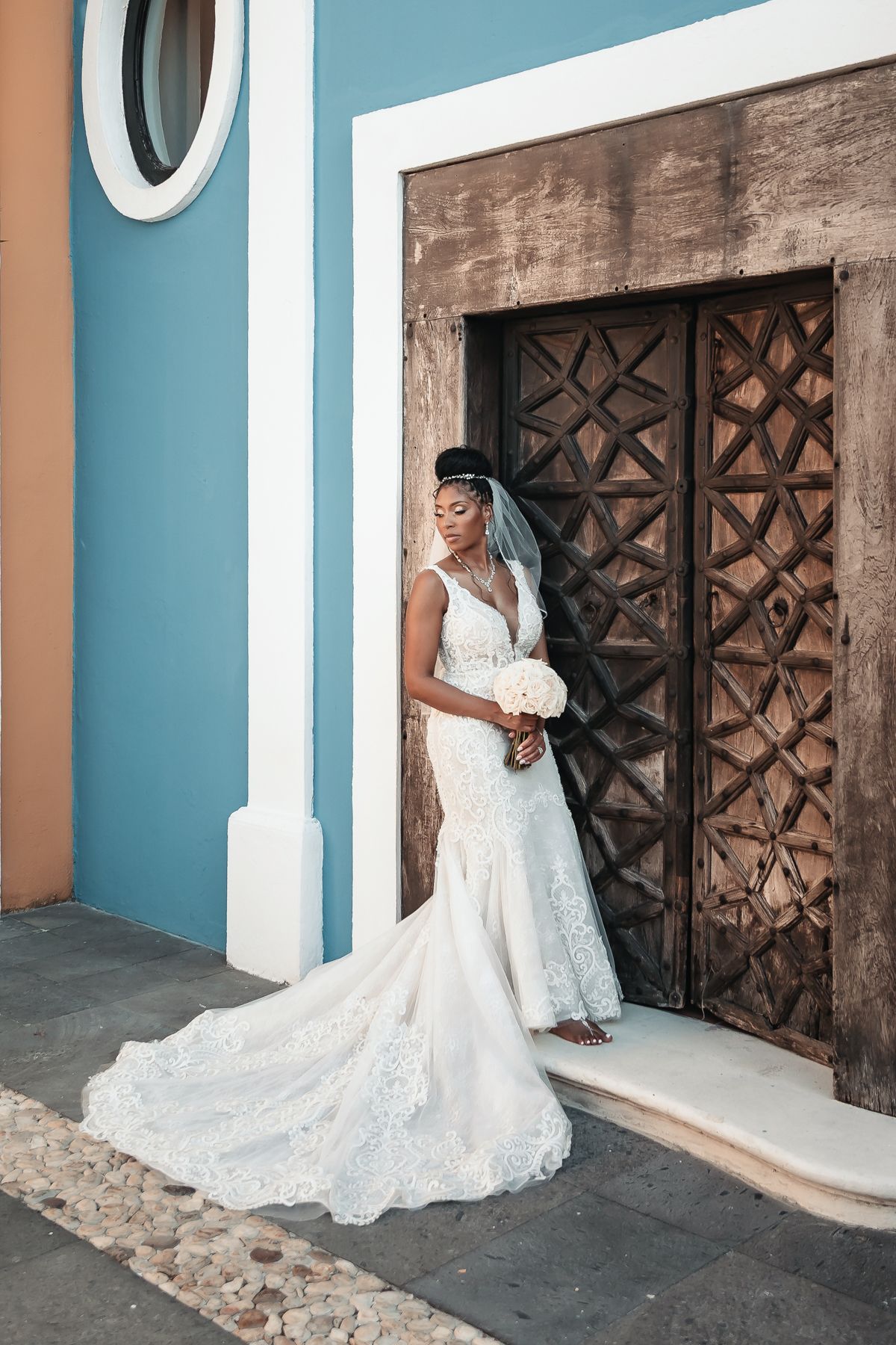Bride in white lace mermaid gown holding bouquet stands before ornate wooden door on cobblestone street.