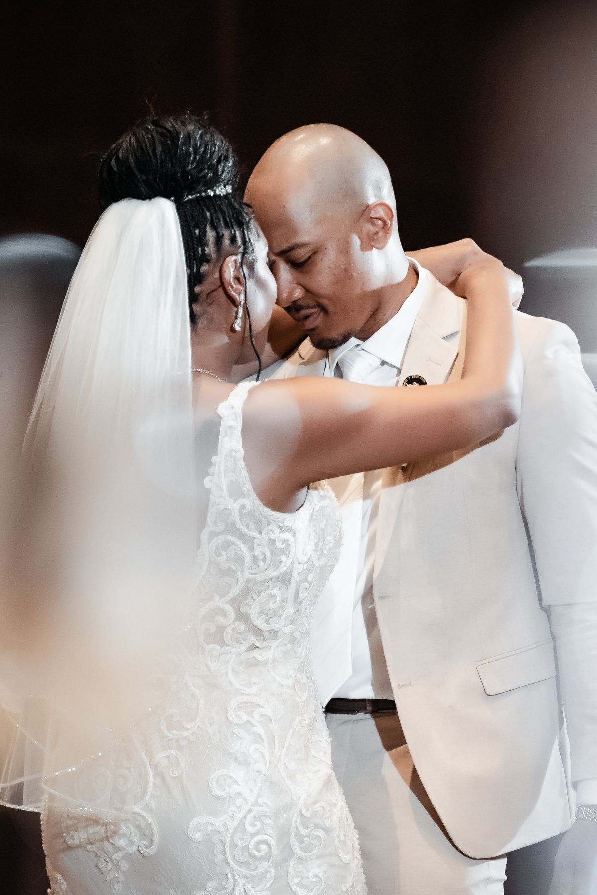 Emotional newlywed couple embracing at wedding ceremony, bride in lace gown with veil, groom in white suit.