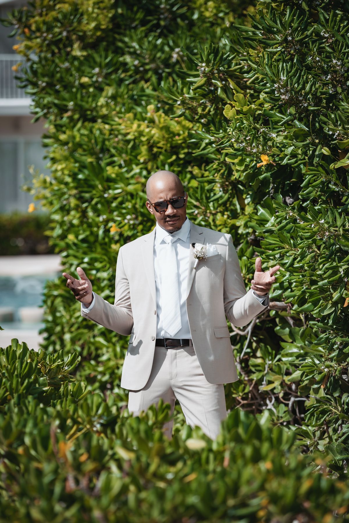 Bald groom in beige suit and sunglasses gestures with hands outdoors near lush green hedges on sunny day.