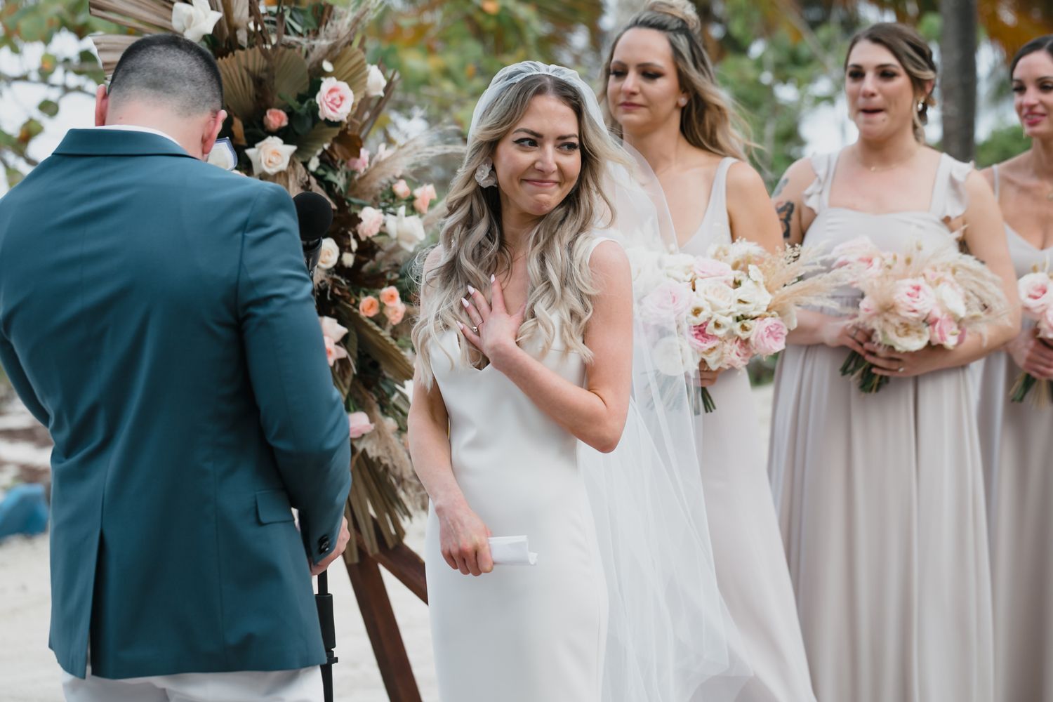 Smiling bride in white gown stands at outdoor wedding altar with bridesmaids holding pink bouquets.