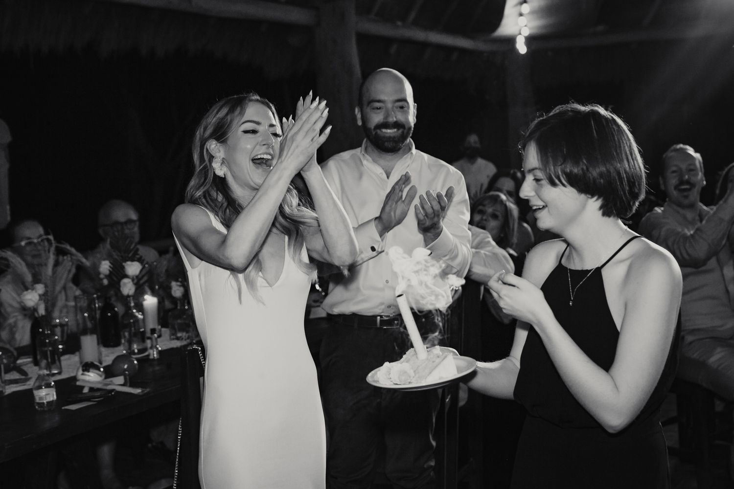 Black and white photo of bride laughing while guests clap around a lit cake at wedding reception.