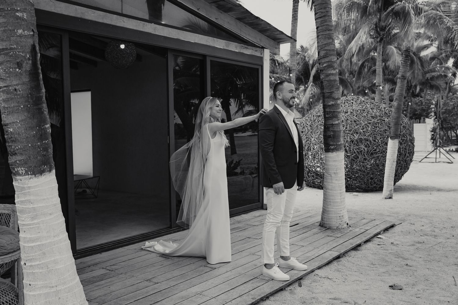 Black and white photo of groom twirling bride in wedding dress and veil outside a beach venue with palm trees.
