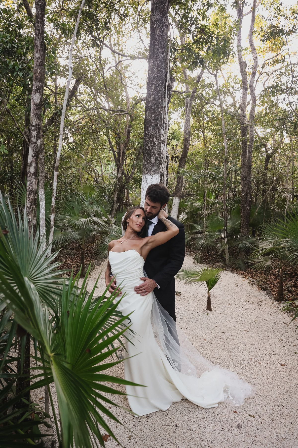 Bride and groom embrace on sandy path surrounded by lush tropical forest in elegant wedding attire.