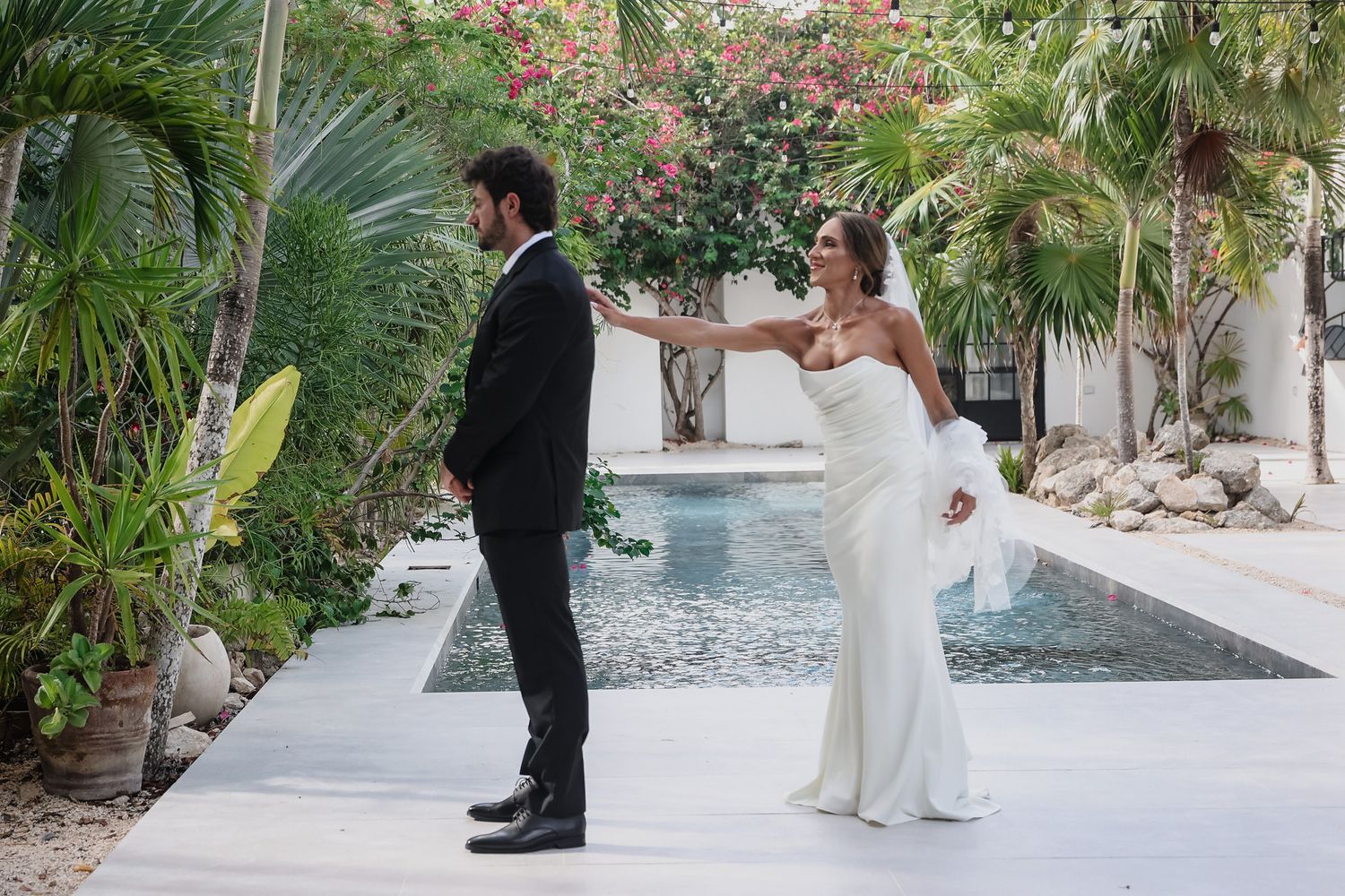 Bride and groom sharing a romantic first look moment by a pool surrounded by tropical flowers and palm trees.