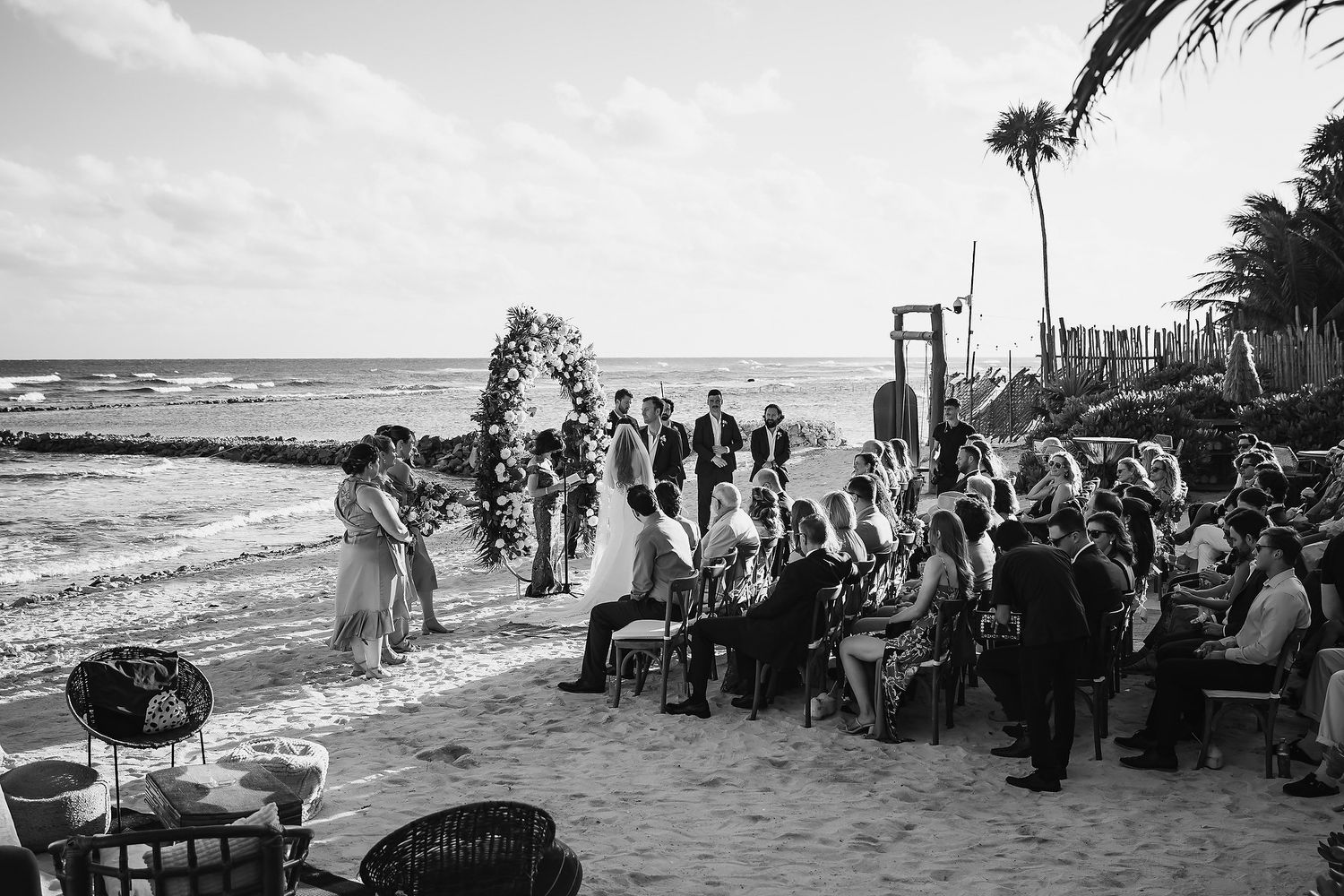 Black and white beach wedding ceremony with guests seated by the ocean, floral arch, and palm trees in background.