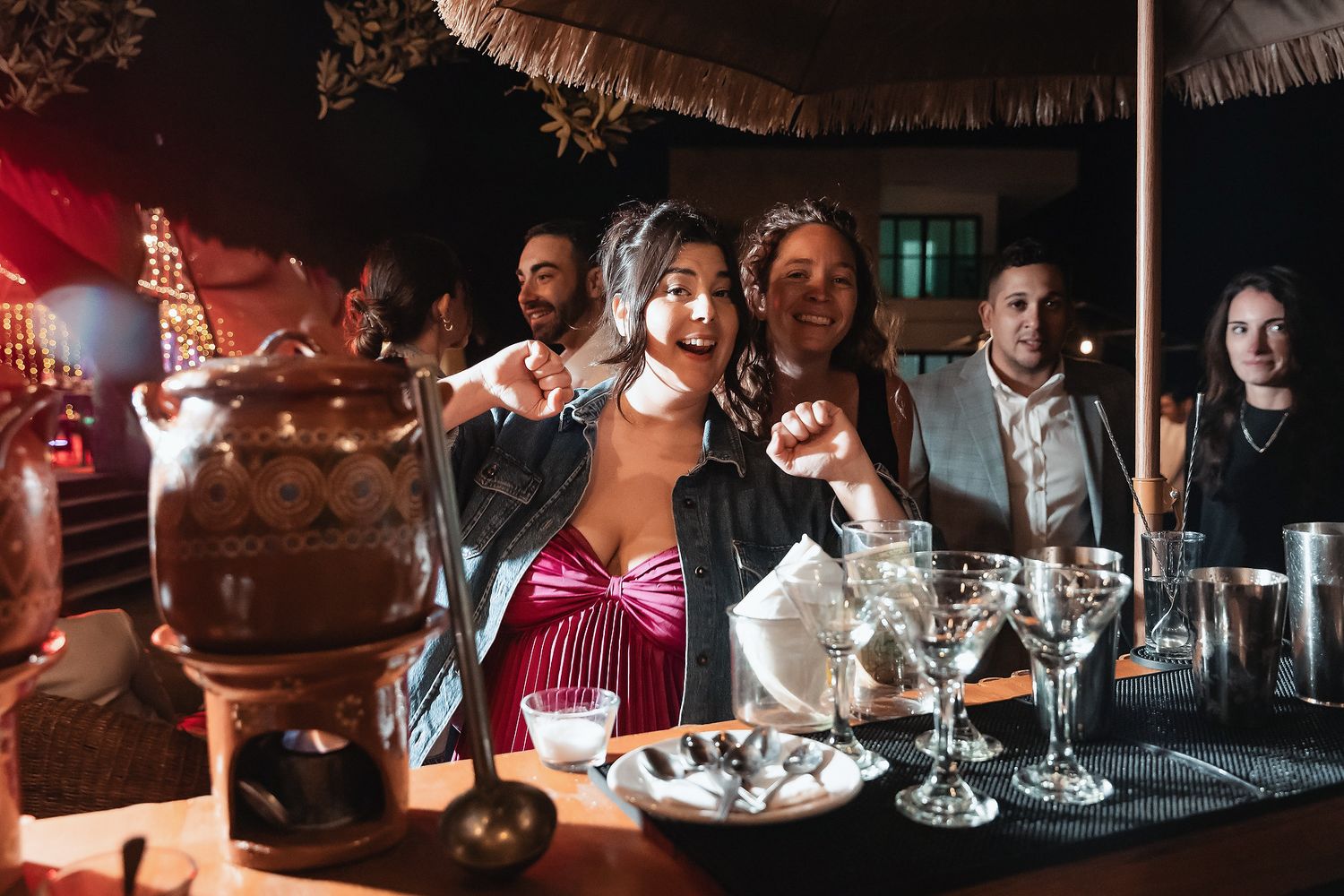 Women celebrating at an outdoor bar at night, with cocktail glasses and festive decorations in background.