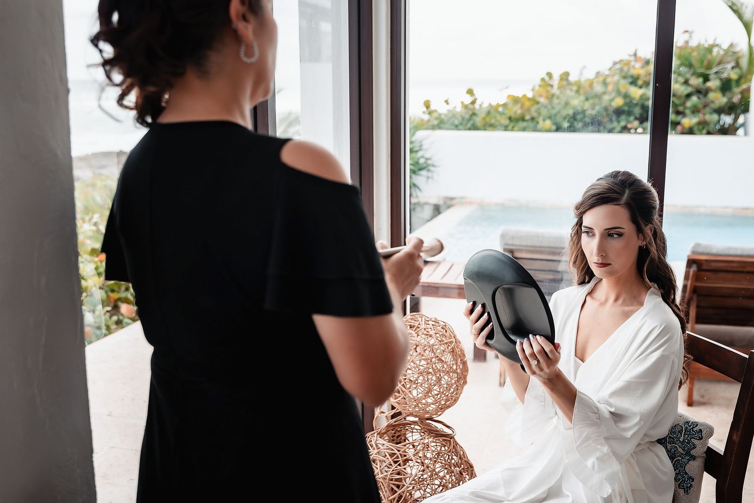 Bride in white robe holds mirror while bridesmaid in black dress assists with wedding preparations indoors.