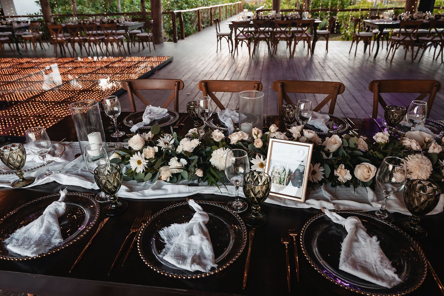 Elegant wedding reception table with floral centerpieces, gold-rimmed chargers, white napkins, and candles on a dark tablecloth.