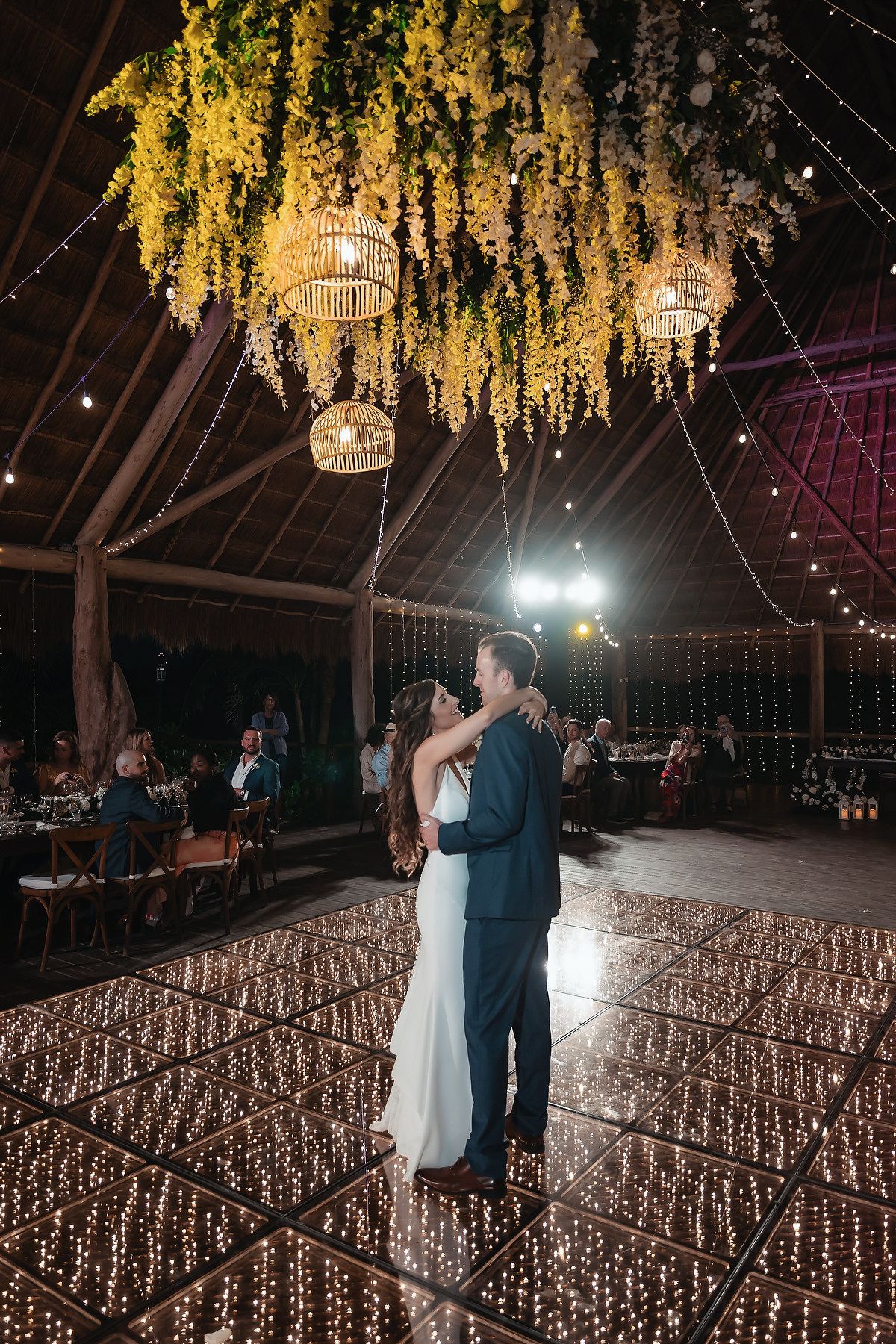 Couple shares first dance on illuminated dance floor under hanging greenery and Edison lights in elegant barn wedding venue.