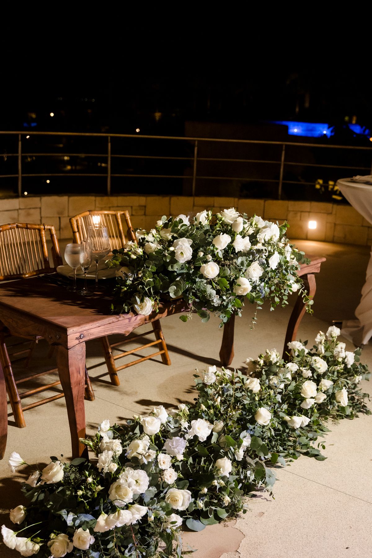 Elegant outdoor wedding reception with white roses, greenery arrangements, and wooden furniture on a terrace at night.