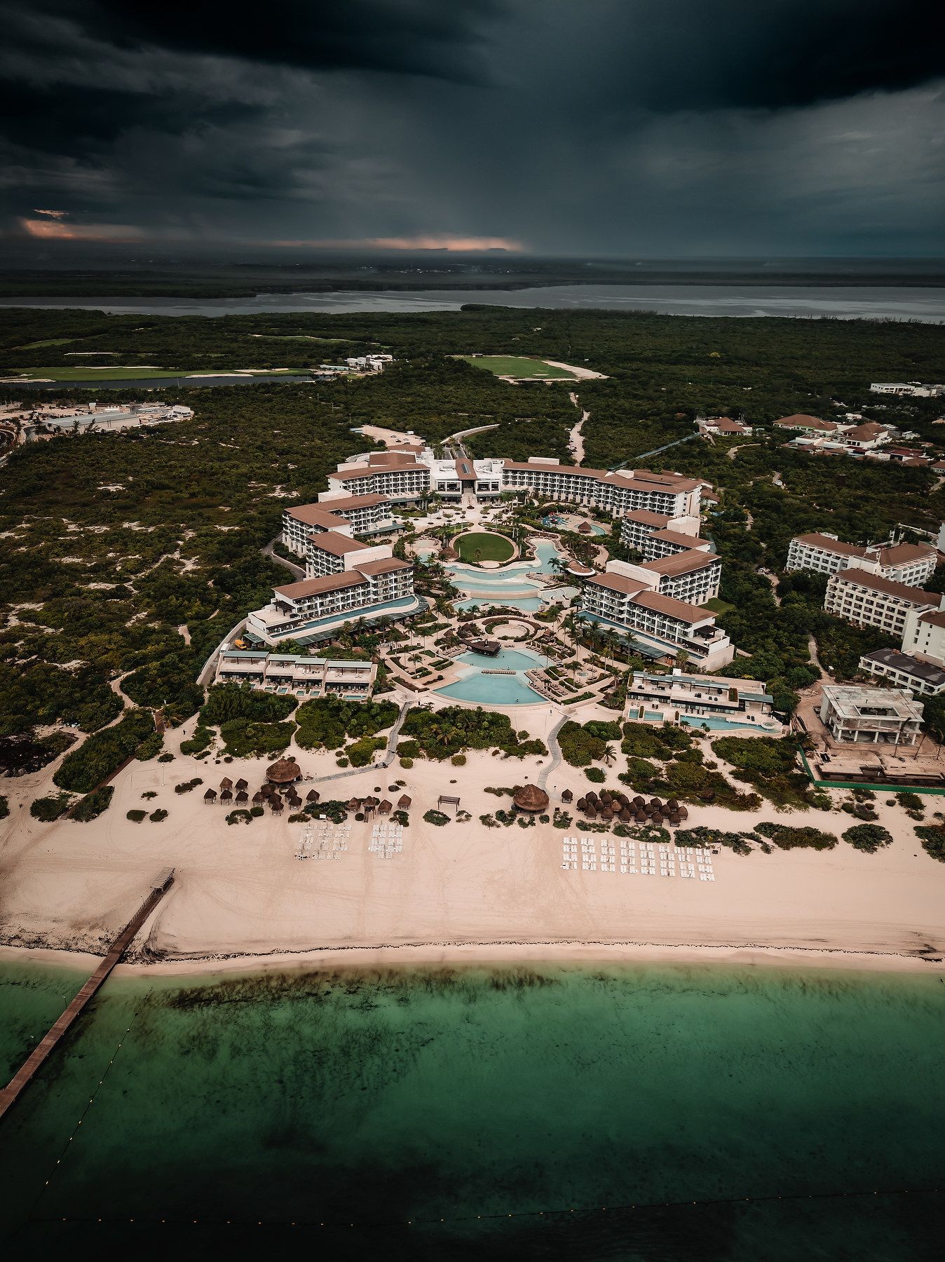 Aerial view of a luxury beachfront resort with pools, surrounded by lush greenery and turquoise waters under stormy skies.