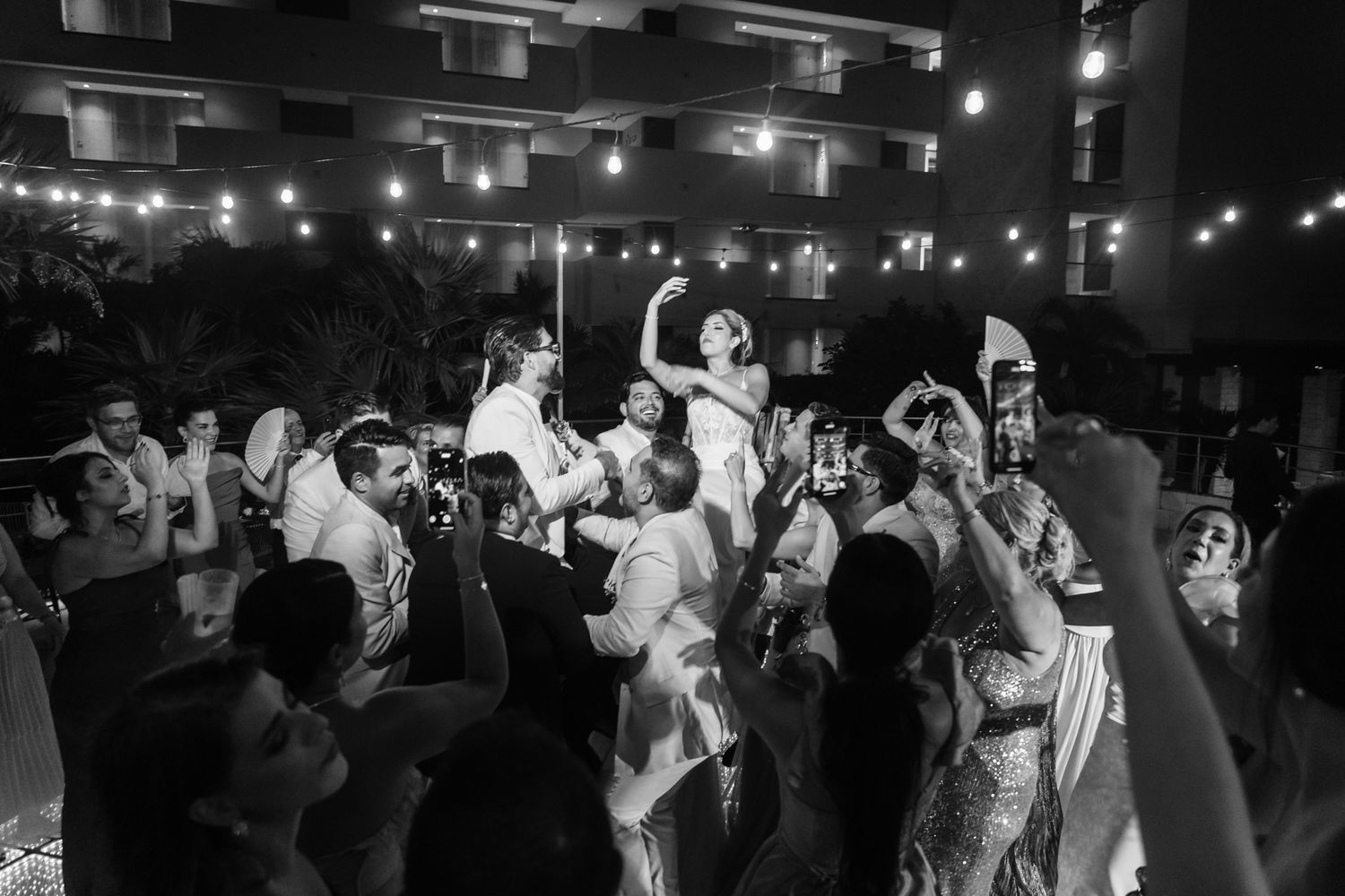 Black and white wedding reception photo of guests lifting bride and groom in celebration under string lights at night.