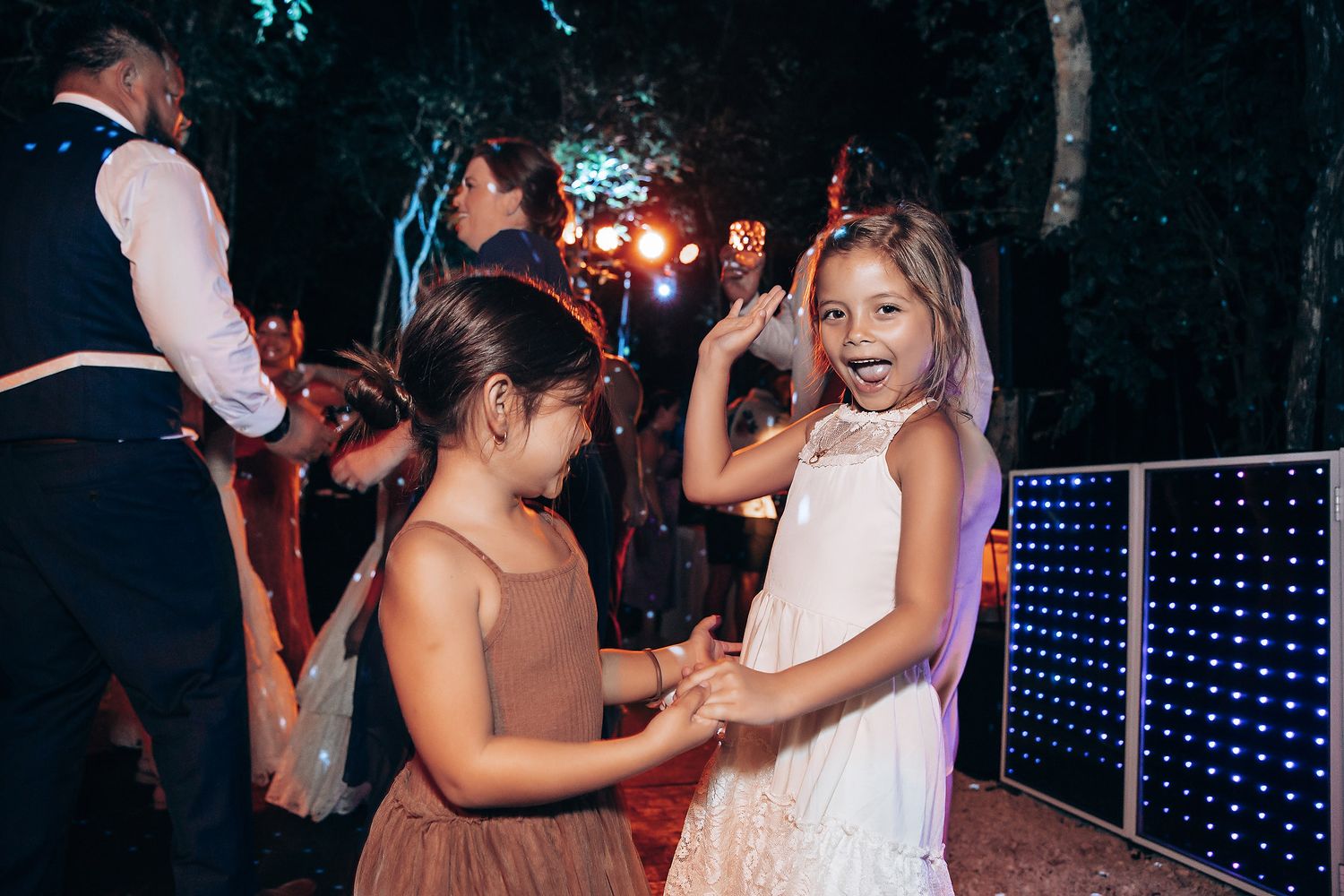 Children in party attire dance together on an outdoor dance floor with string lights in the background.