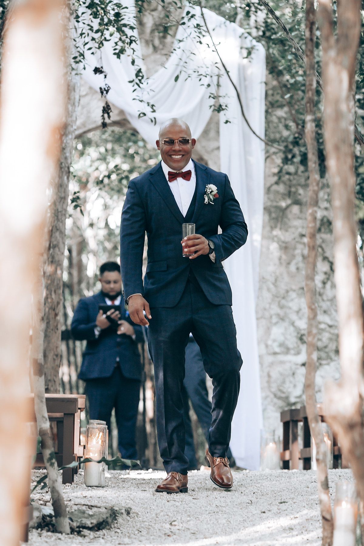 Groomsmen in navy suits walking down a tree-lined path during an outdoor wedding celebration.