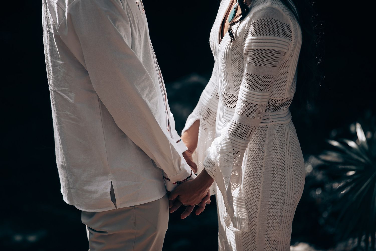 Couple in white outfits holding hands during an intimate wedding ceremony in a natural setting.