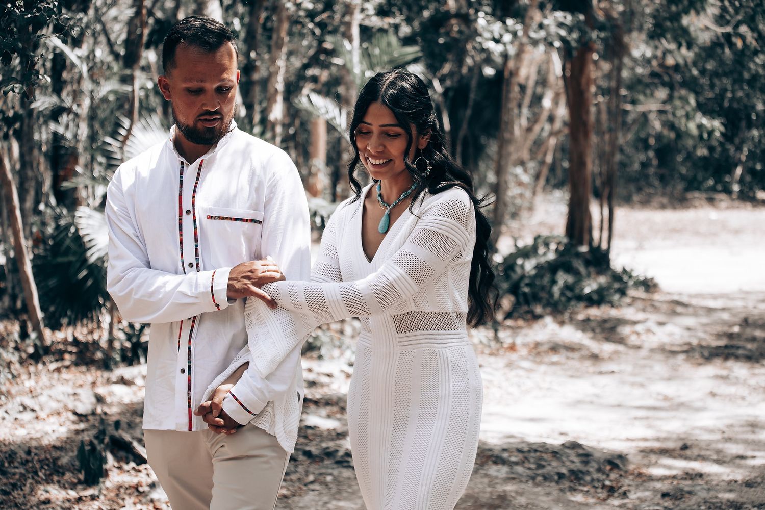 A couple in white outfits share a romantic moment together in a sunlit wooded area.