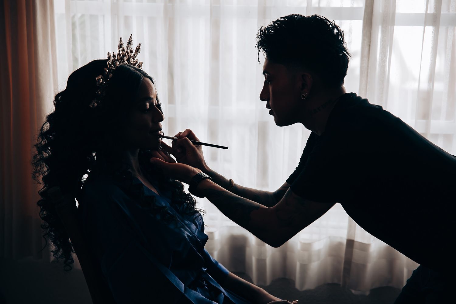 Silhouette of makeup artist applying cosmetics on bride by window.