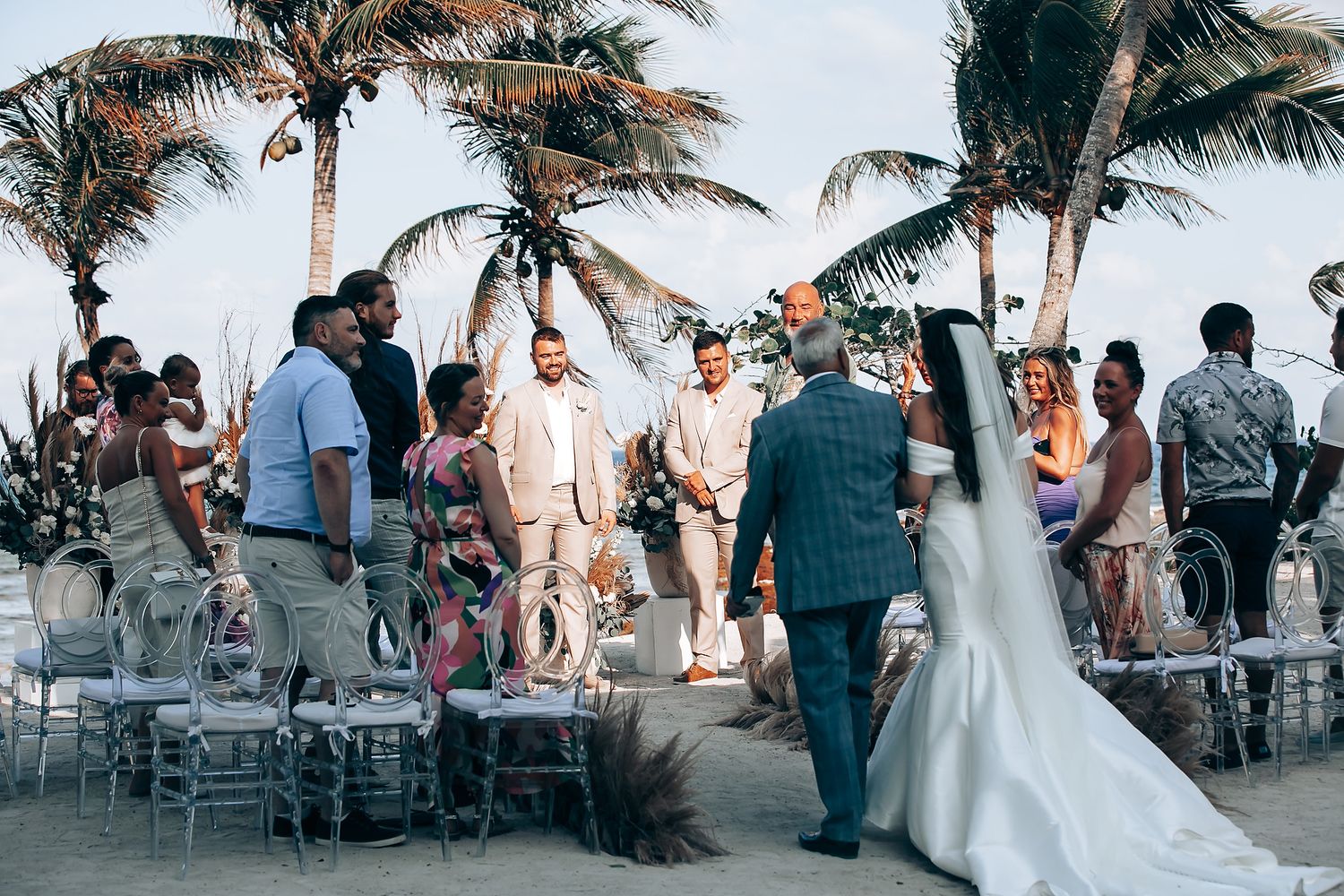 Wedding guests celebrate on a tropical beach at sunset with palm trees and white chairs arranged for the ceremony.
