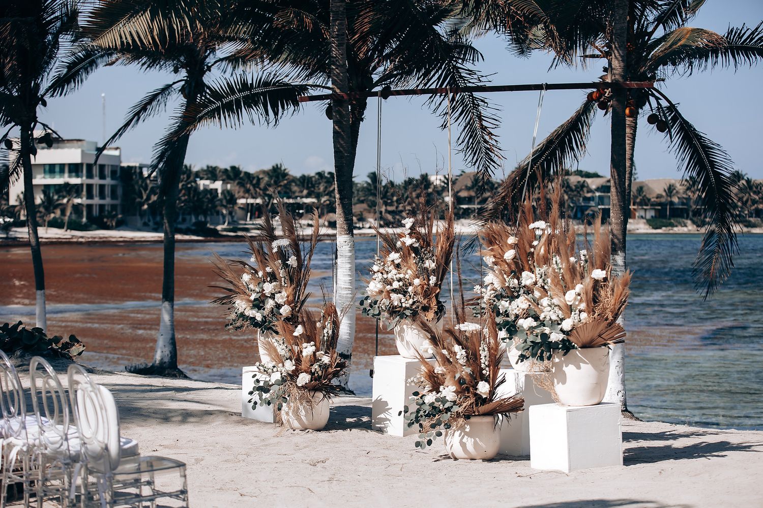 A beachside wedding ceremony setup with white chairs and floral arrangements under palm trees by the ocean.
