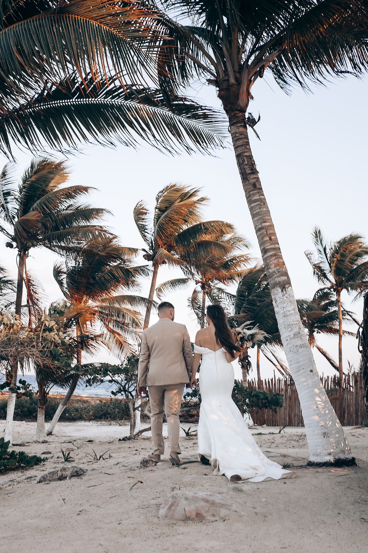 A couple shares a romantic moment on a windy beach with swaying palm trees during a tropical wedding photoshoot.