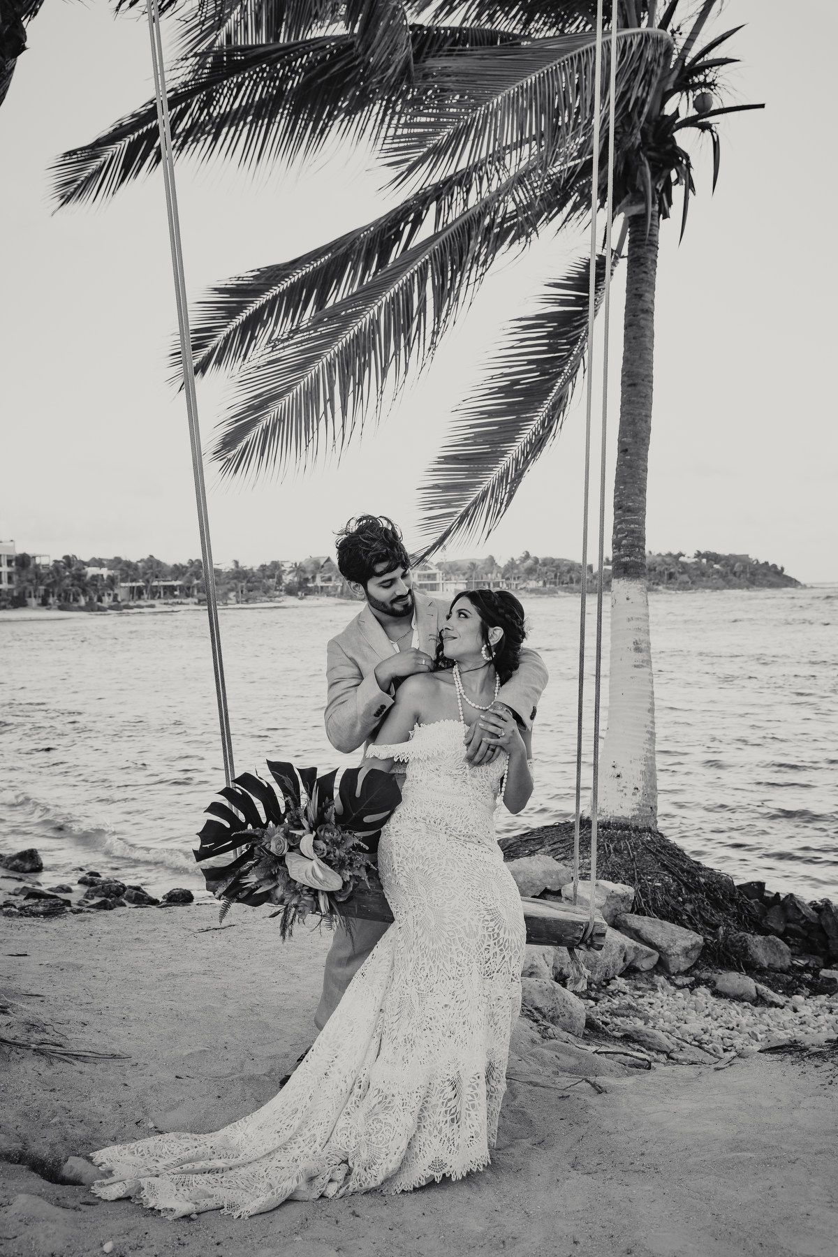 Black and white photo of bride and groom on beach swing under palm tree.