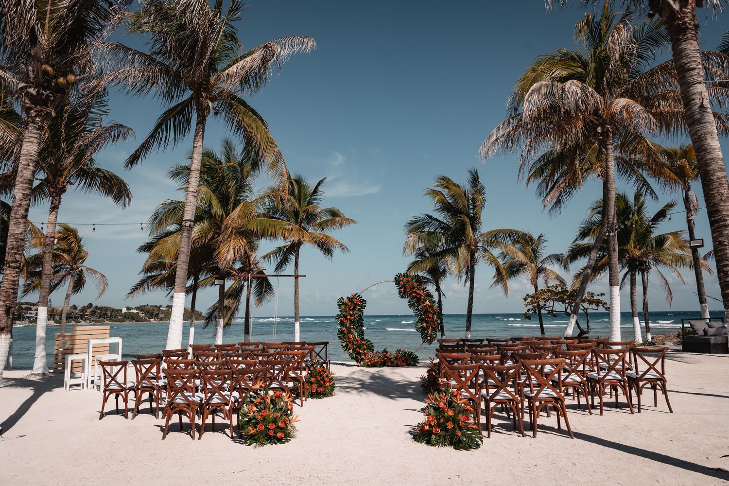 Beach wedding ceremony setup with wooden chairs, floral arch, and palm trees overlooking turquoise ocean waters.