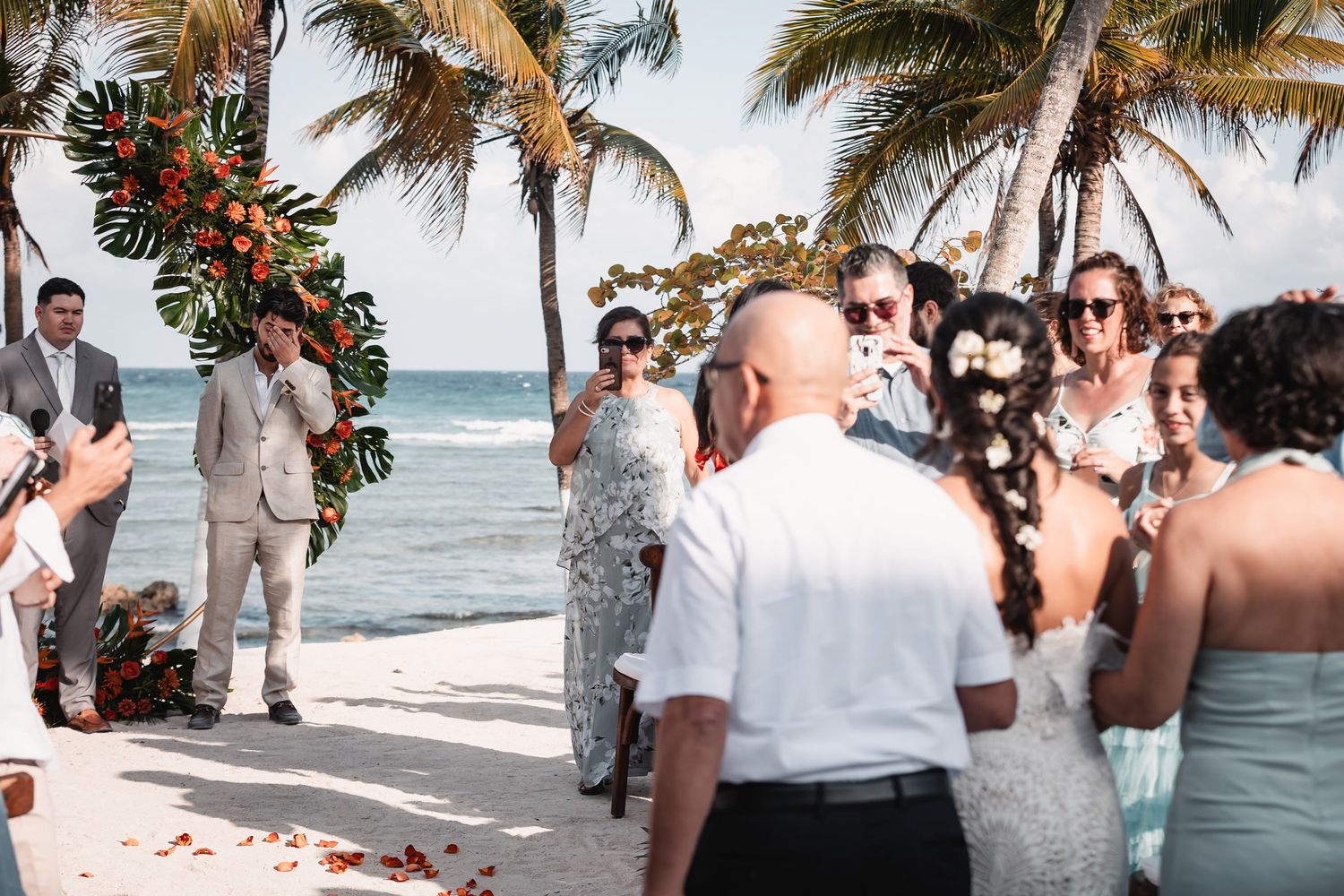 Beach wedding ceremony with guests watching couple at floral arch by ocean with palm trees.