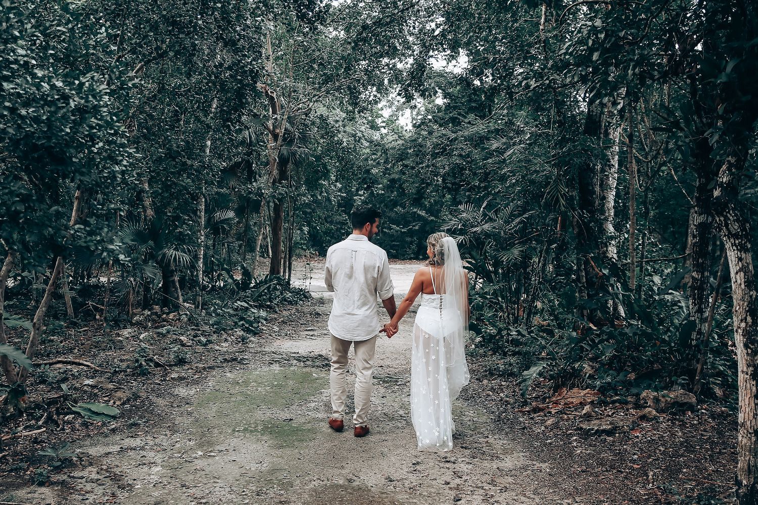 A couple walks hand in hand through a serene forest path in wedding attire.