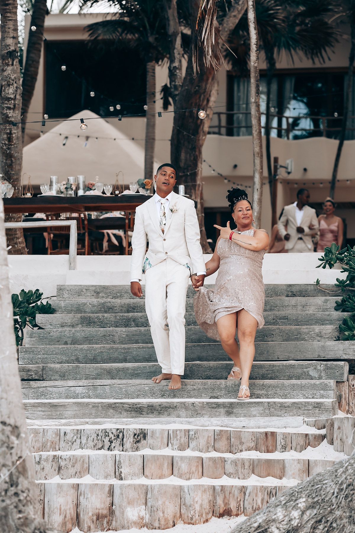 A couple in white attire poses on stone steps near a beach for a casual photo session.