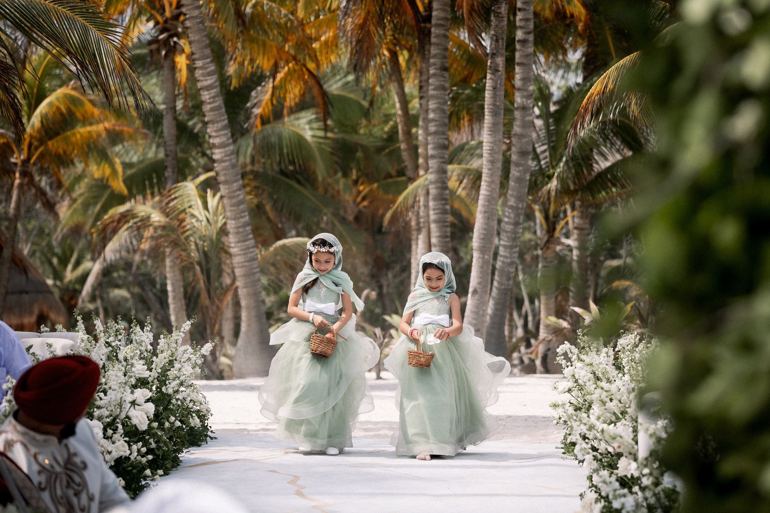 Two flower girls in mint green gowns walking down tropical palm-lined wedding aisle.