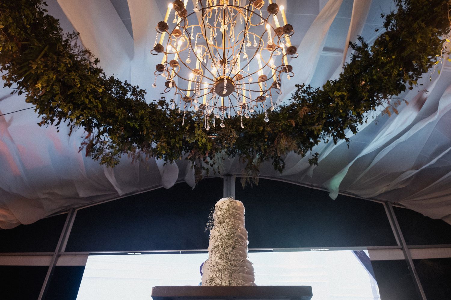 Elegant tiered white wedding cake displayed beneath chandelier draped with lush greenery.