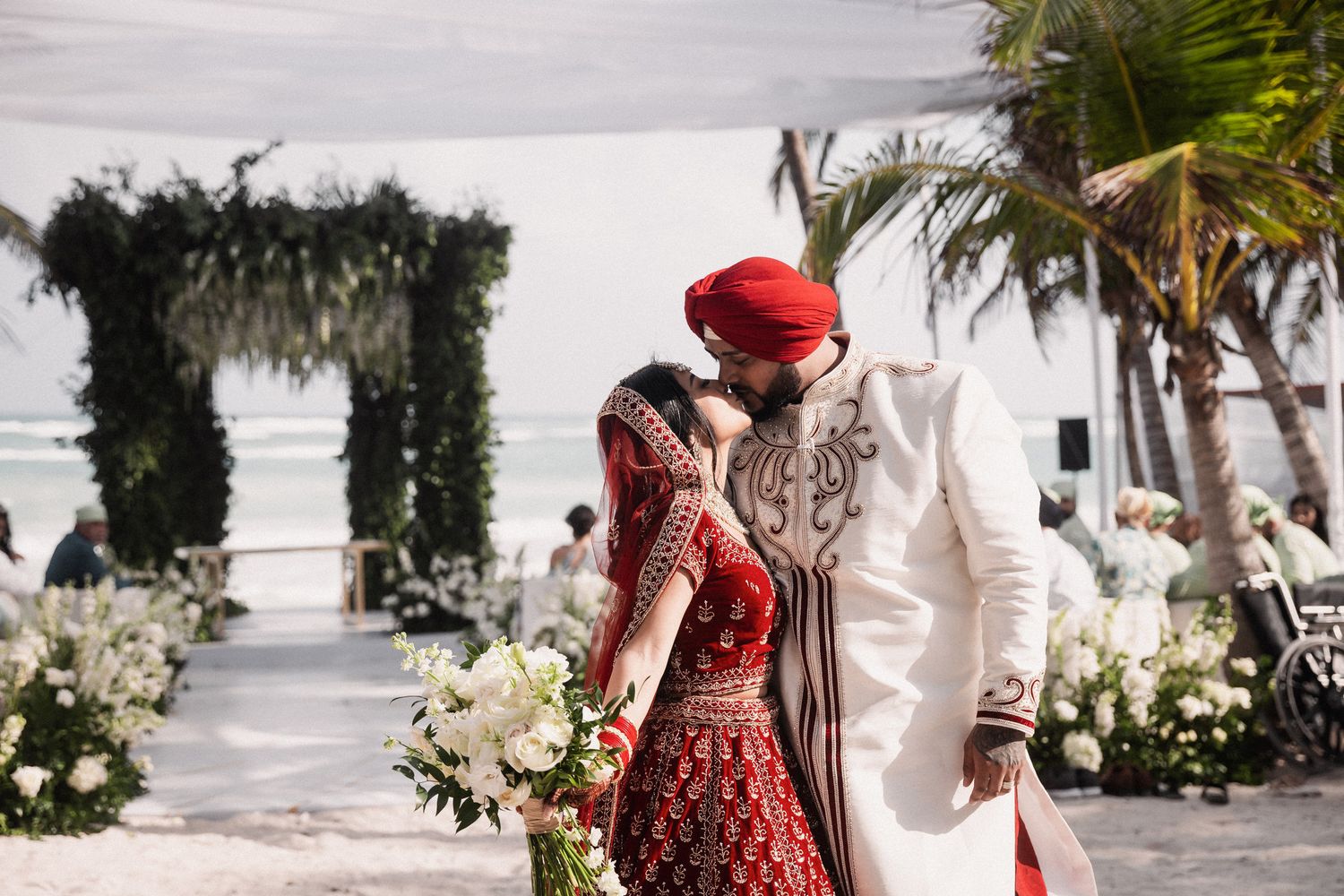 Indian bride and groom sharing kiss at tropical beach wedding ceremony with floral arch backdrop.