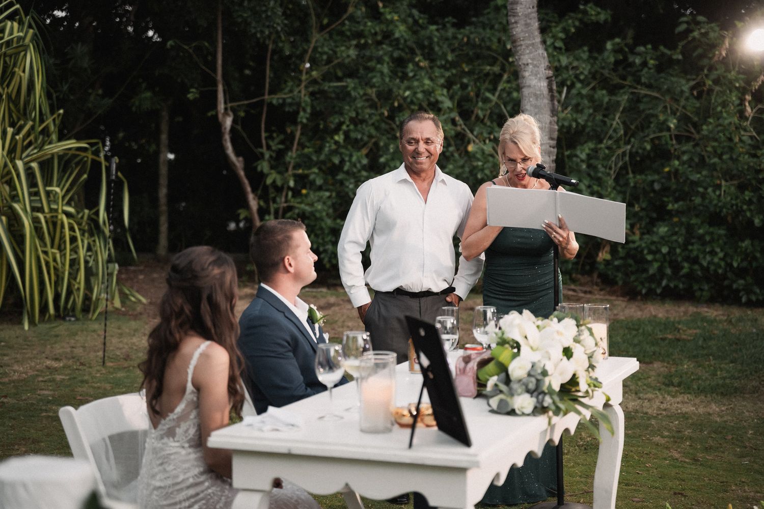 Wedding guests laugh during outdoor reception speech at elegantly decorated white table surrounded by tropical greenery at night.