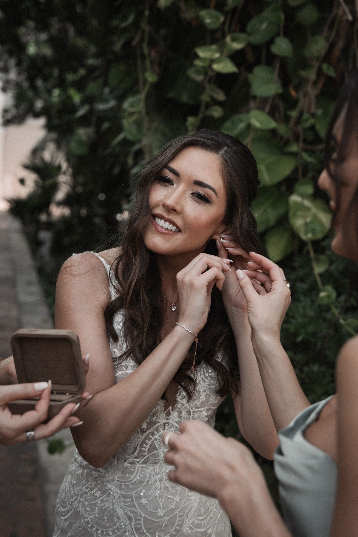 Smiling bride puts on earring with help from bridesmaid surrounded by lush green ivy before wedding ceremony.