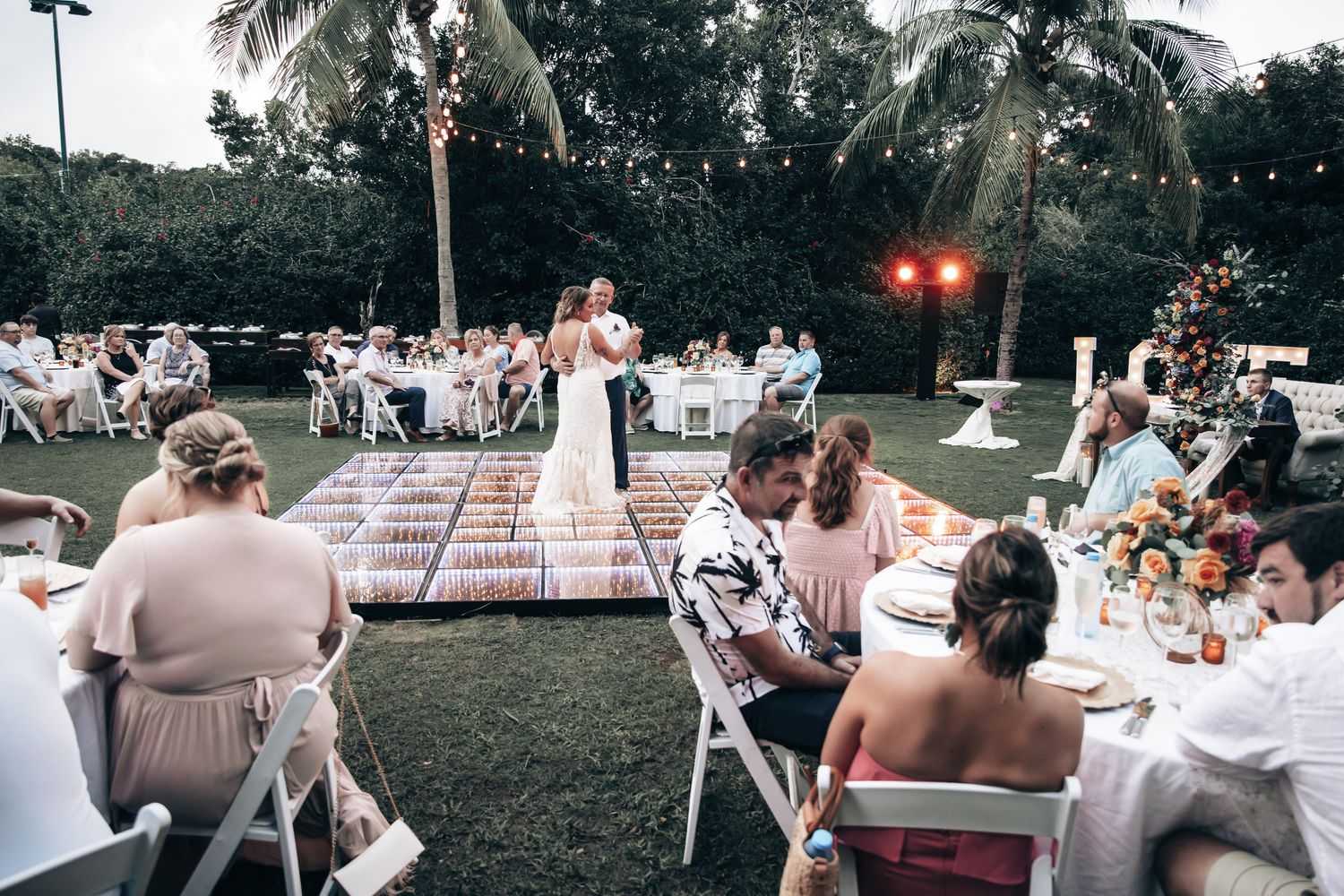 Outdoor tropical wedding reception with guests seated at decorated tables while couple stands on dance floor.