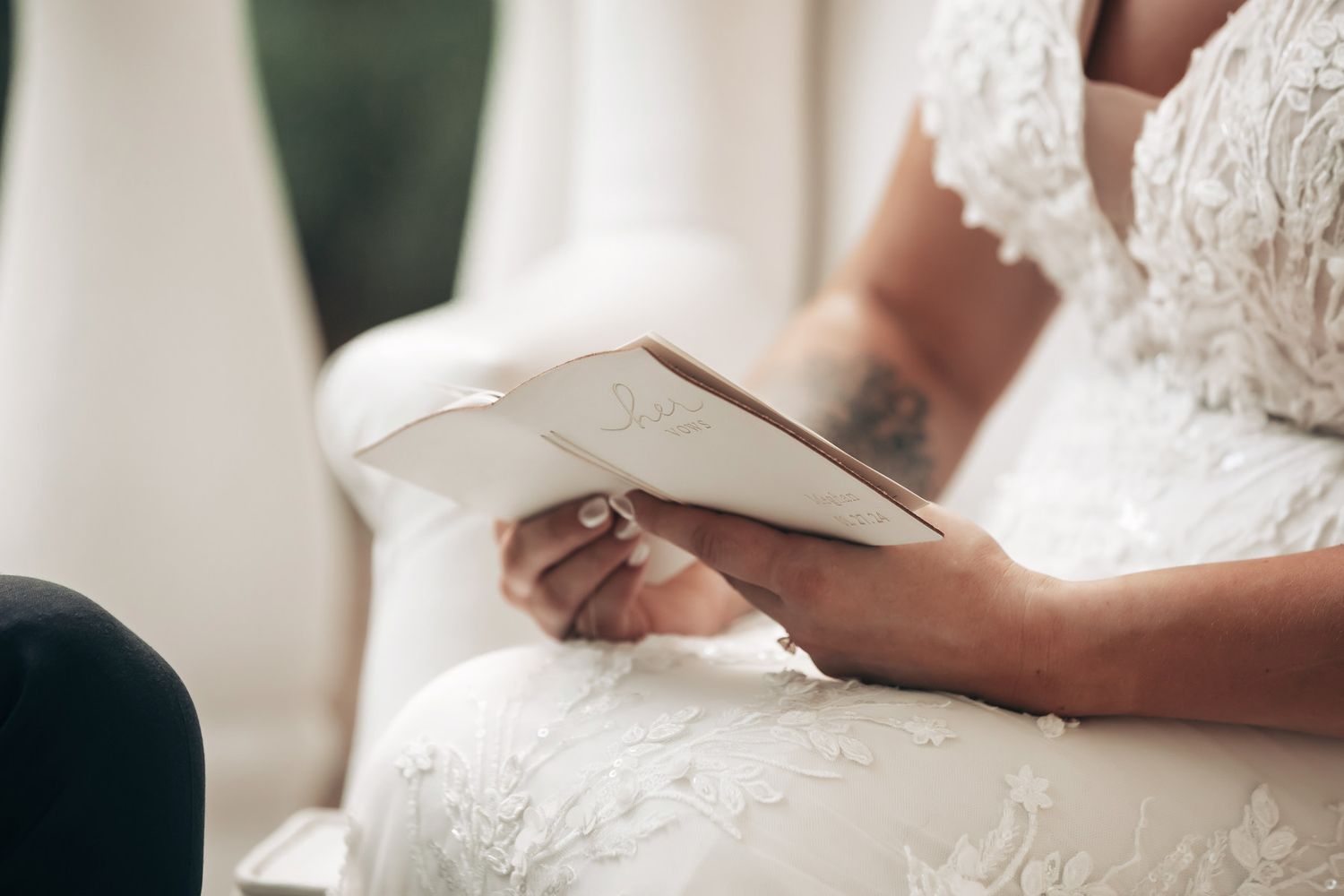 Bride in lace wedding dress reading vows from a small book while seated, showing tattooed arm.