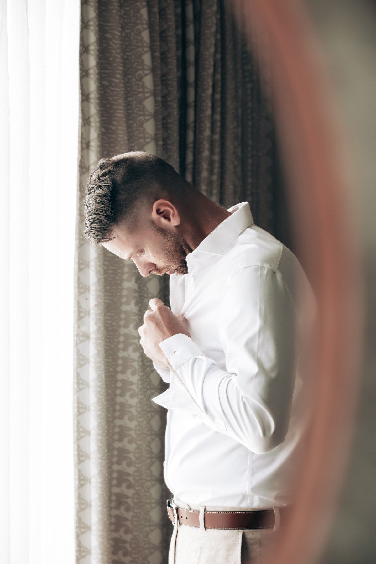 Groom buttoning white dress shirt while standing by a curtained window.