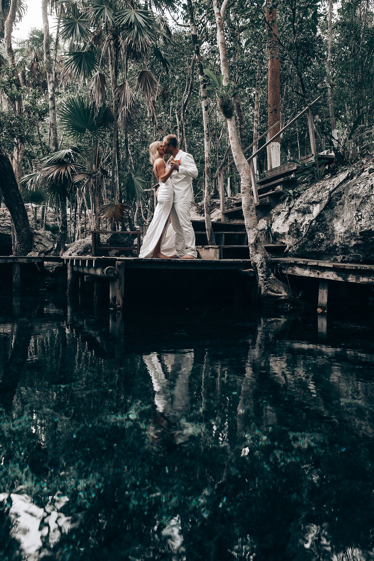 A couple shares a romantic moment on a wooden boardwalk over dark water surrounded by trees.