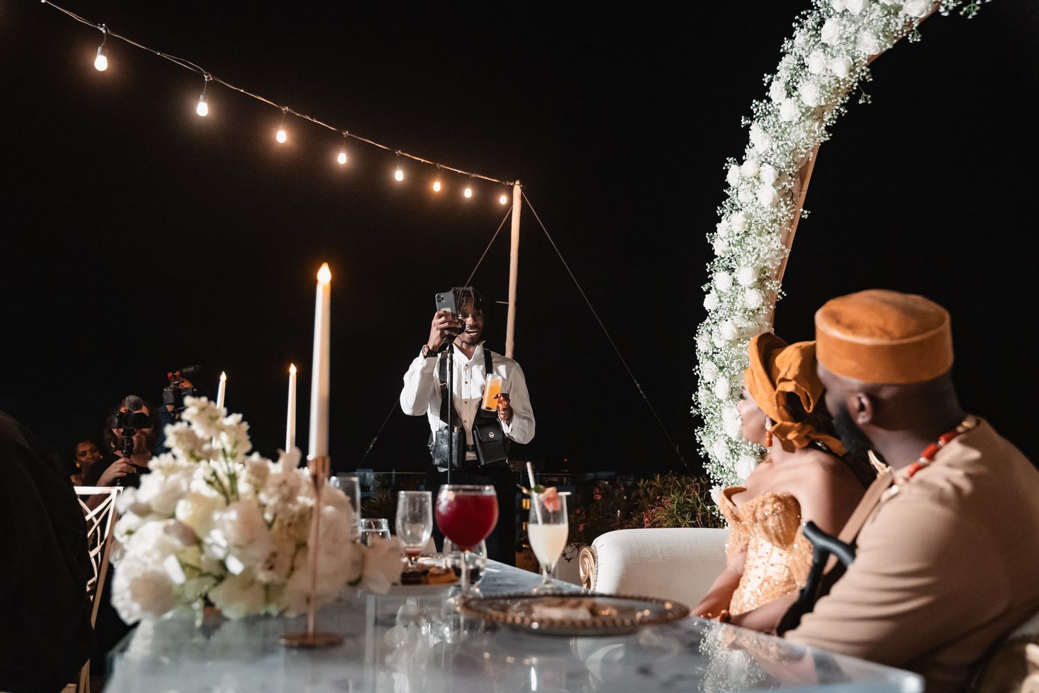 Wedding guest giving speech at outdoor nighttime reception with floral arch and string lights.