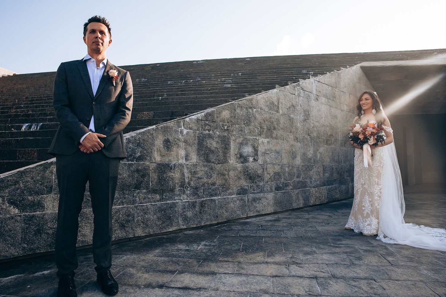 A wedding couple poses in front of a rustic stone wall with dramatic lighting creating romantic silhouettes.