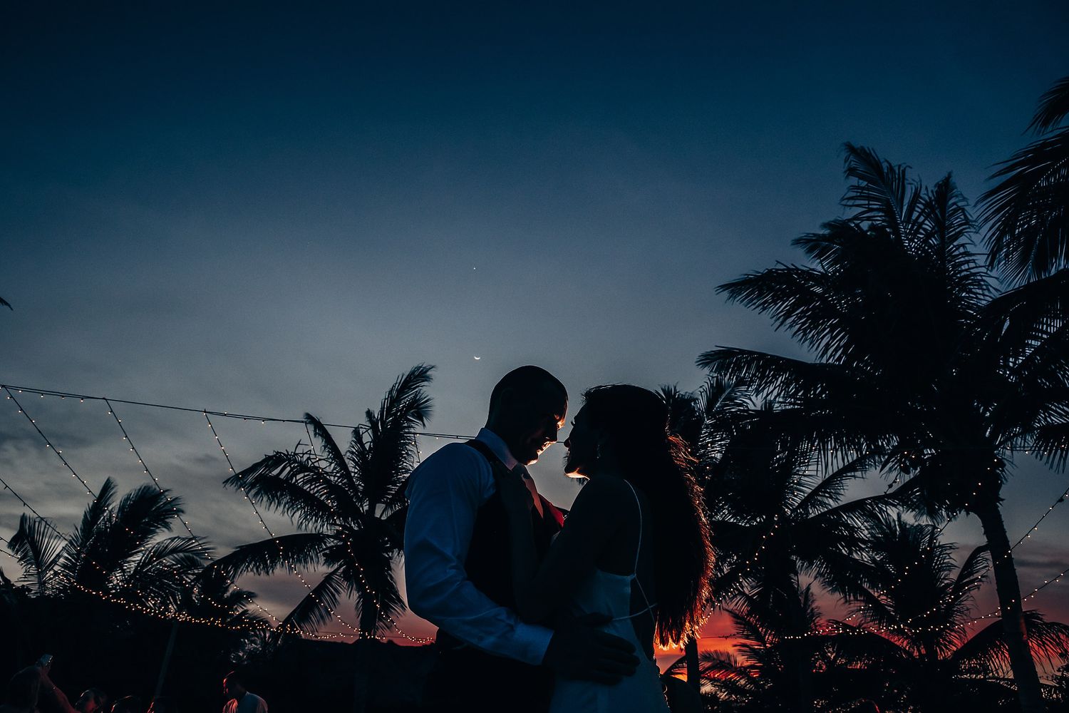 Silhouettes of palm trees against a dramatic sunset sky at a beach wedding.