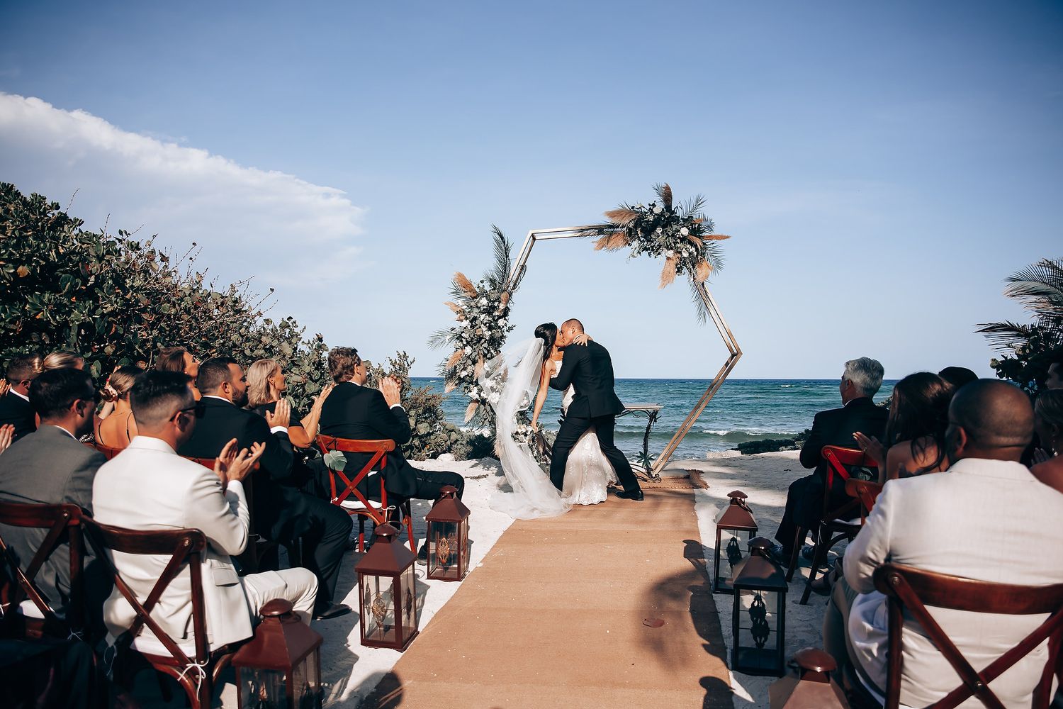 Seaside wedding celebration with guests gathered around a decorated wooden arch by the ocean.