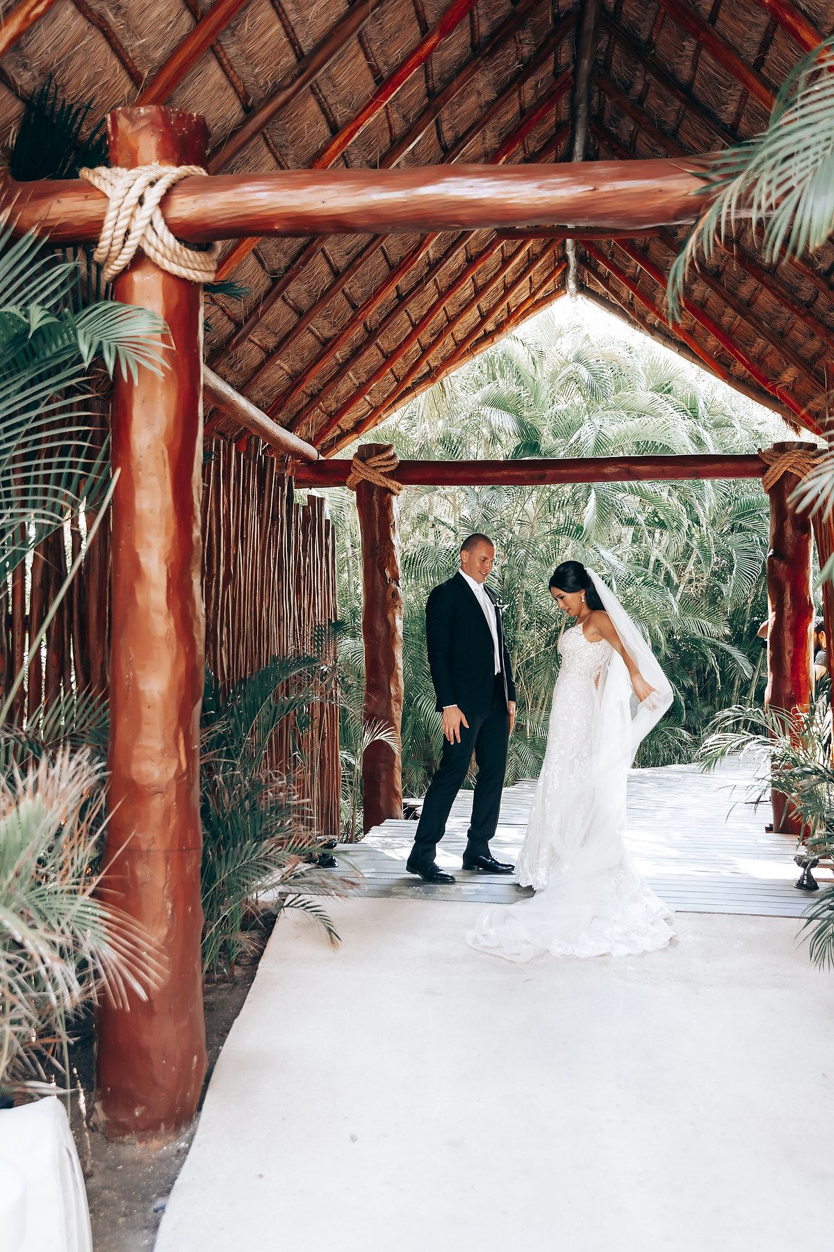 A couple in wedding attire poses under a rustic wooden pavilion with palm trees visible in the background.