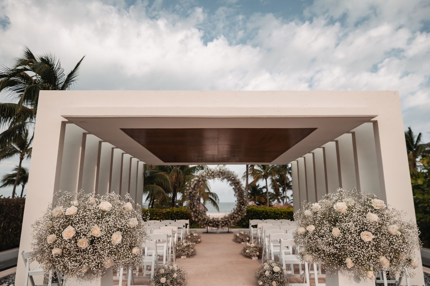 Elegant outdoor wedding ceremony setup with floral arch, white chairs, and tropical palm trees.