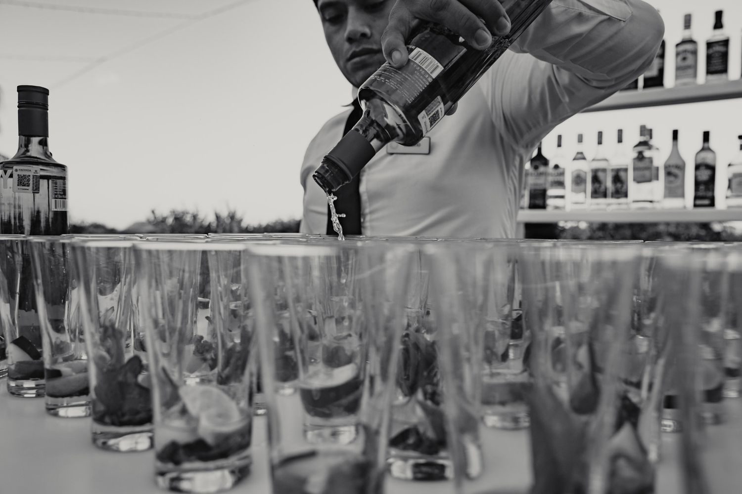 Bartender pouring drinks into a row of shot glasses at a wedding bar setup in black and white.