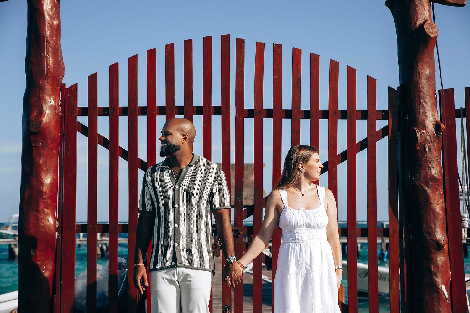 Couple holding hands in front of a red wooden gate by the ocean on a sunny day.