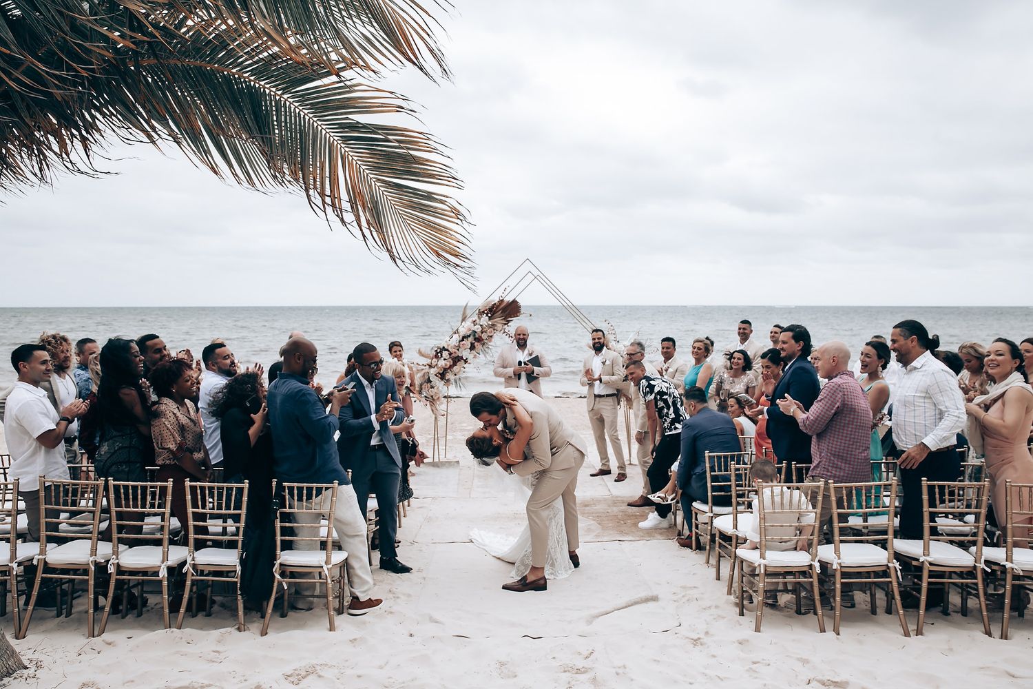 Panoramic view of beachside wedding ceremony with guests seated on white chairs under palm fronds.