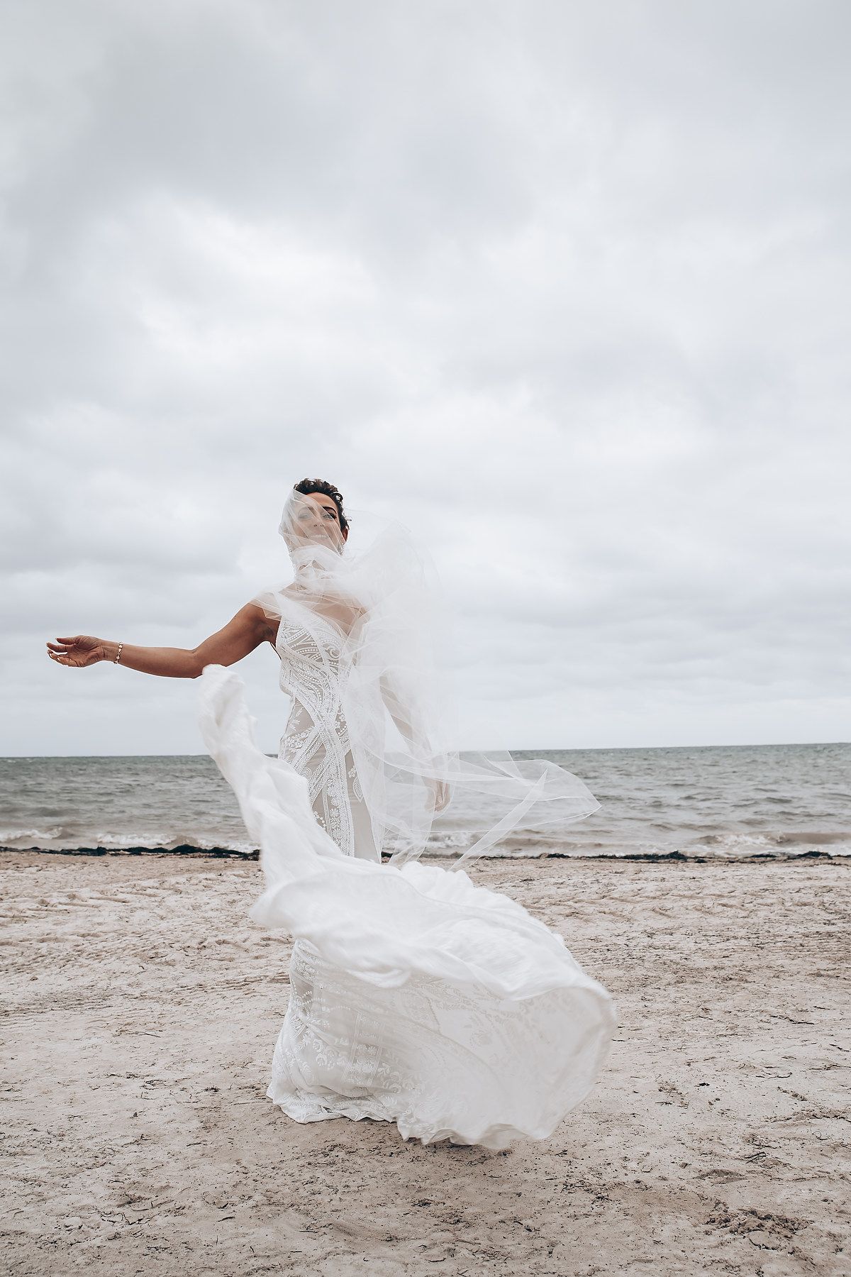 White wedding dress flowing dramatically on a cloudy beach with waves in the background.