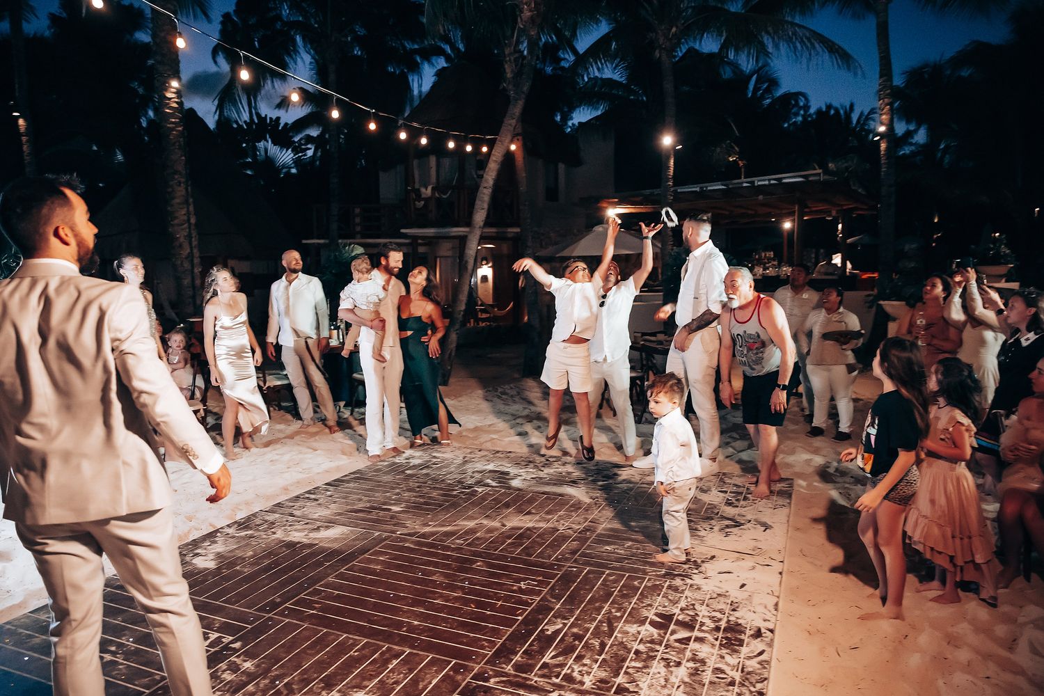 Wedding party dancing on a beachfront deck illuminated by string lights at dusk.