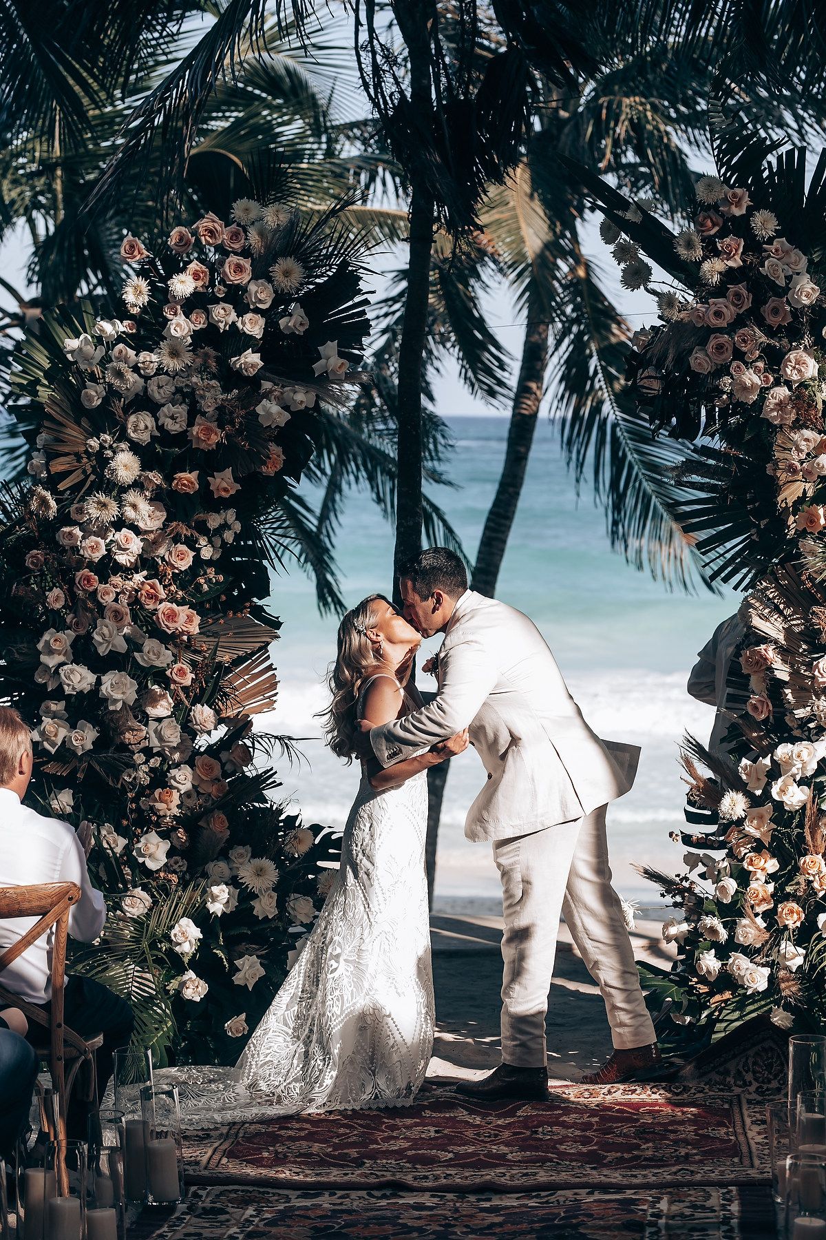 Beachside wedding ceremony setup with floral arch, palm trees, and ocean views.